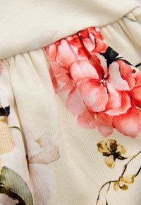 Blomstret stof i bløde creme- og lyserøde toner med ribbet tekstur og opsamlet design ved taljen, der fremhæver elegante blomsterdetaljer.