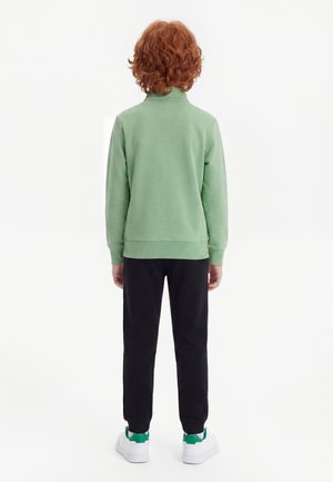 Barn med krøllet rødt hår står med ryggen til iført en lysegrøn sweatshirt, sorte bukser og hvide sneakers med grønne accenter.