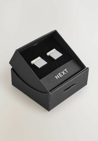 Stříbrné kovové manžetové knoflíčky s obdélníkovým tvarem, se lesklým povrchem a jemným horizontálním pruhovaným akcentem, prezentované v černé krabičce.