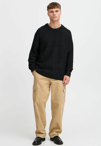Pulover negru din tricot cu decolteu rotund și margini ondulate, asortat cu pantaloni cargo bej cu buzunare laterale și croială relaxată. Pantofi negri.
