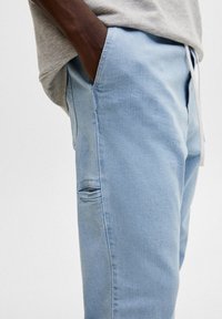 Pantalon en denim bleu clair à coupe décontractée, avec une poche latérale et une taille à cordon, présenté sur un fond neutre.