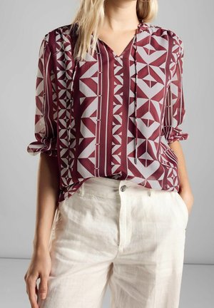 Kobieta w bordowo-białej bluzce z geometrycznym wzorem, z podwiniętymi rękawami i białych spodniach z wysokim stanem, z ręką w kieszeni.