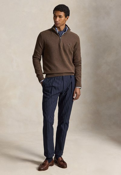Άνδρας που φοράει καφέ πουλόβερ με φερμουάρ μέχρι τη μέση, μπλε φουλάρι με πουά, μπλε παντελόνι με ρίγες και καφέ μοκασίνια με φούντες, στέκεται μπροστά σε ουδέτερο φόντο.