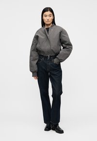 Γυναίκα στέκεται φορώντας γκρι μπουφάν bomber, σκούρα ίσια τζιν παντελόνια και μαύρα μποτάκια μέχρι τον αστράγαλο μπροστά σε απλό φόντο.