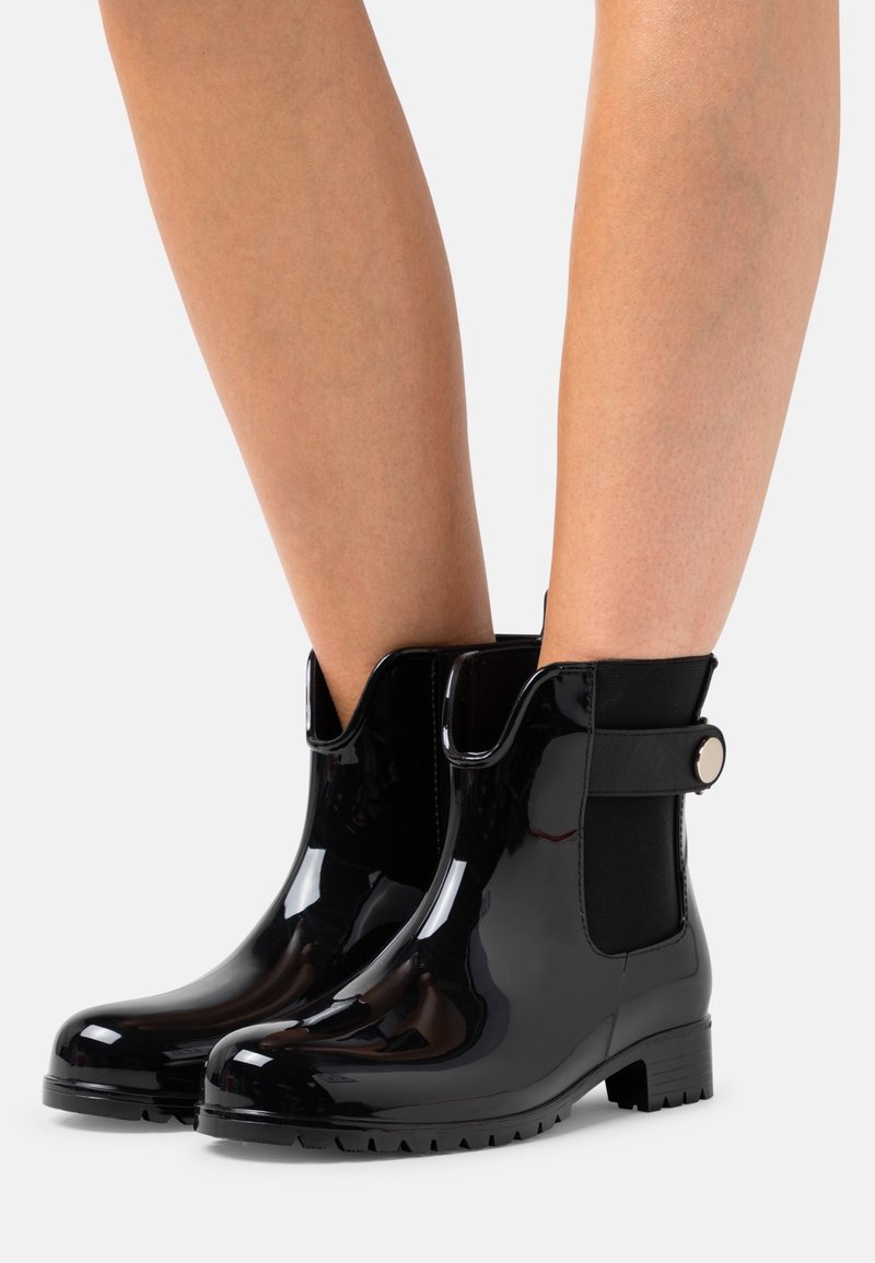 Bottines noires brillantes avec panneaux latéraux élastiques et brides boutonnées portées sur des jambes nues sur fond blanc.