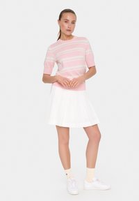 Suéter rosa de manga corta con rayas blancas, combinado con una falda blanca con pliegues. La modelo lleva zapatillas blancas y calcetines claros. Diseño simple y limpio.