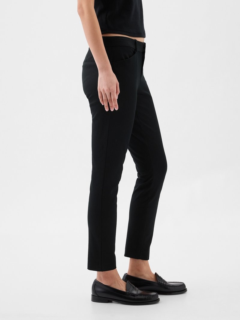Femme portant un pantalon noir ajusté, des mocassins noirs et un haut court noir, se tenant de profil contre un fond clair uni.