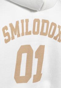 Bílá mikina s kapucí, na které je velký krémový text "SMILODOX" nad "01." Tkanina vypadá měkce a má hladkou texturu.