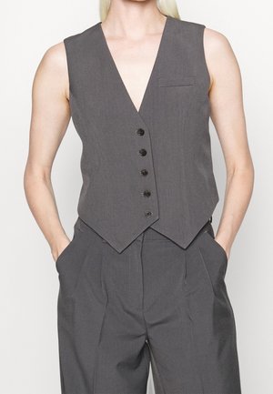 Person iført ærmeløs mørkegrå vest med knapper og matchende plisserede bukser med hænderne i lommerne mod hvid baggrund.