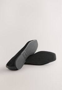 Paire de chaussures plates noires avec semelles en caoutchouc texturées et bouts carrés, positionnées pour montrer à la fois la vue de dessus et de dessous sur un fond uni.