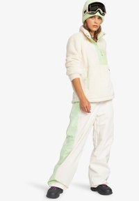 Cream-colored fleece pullover with mint green accents, front pockets, and high collar. White ski pants feature mint side panels. Black winter boots.