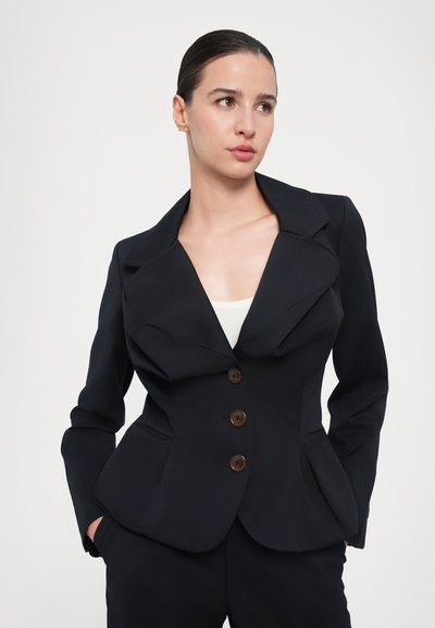Marineblazer med spids krave, fire brune knapper, skrå lommer og peplum-silhuet. Fremstillet af glat, skræddersyet stof.