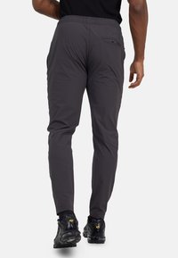 Achteraanzicht van een persoon in donkergrijze slim-fit broek met achterzak en zwarte sneakers met zichtbaar merklogo.