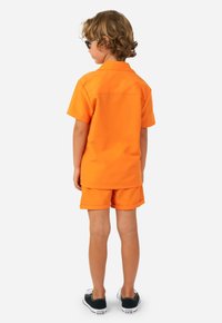 Camisa de manga corta naranja y pantalones cortos a juego, fabricados en tejido suave. Con un yugo en la parte posterior, combinados con zapatillas negras.