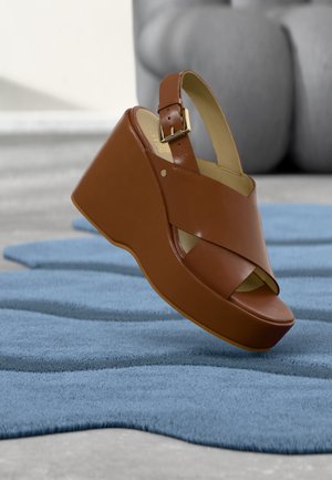 Brun læder kile sandal med justerbar spænde stropp, der hviler på tekstureret blåt tæppe indendørs.