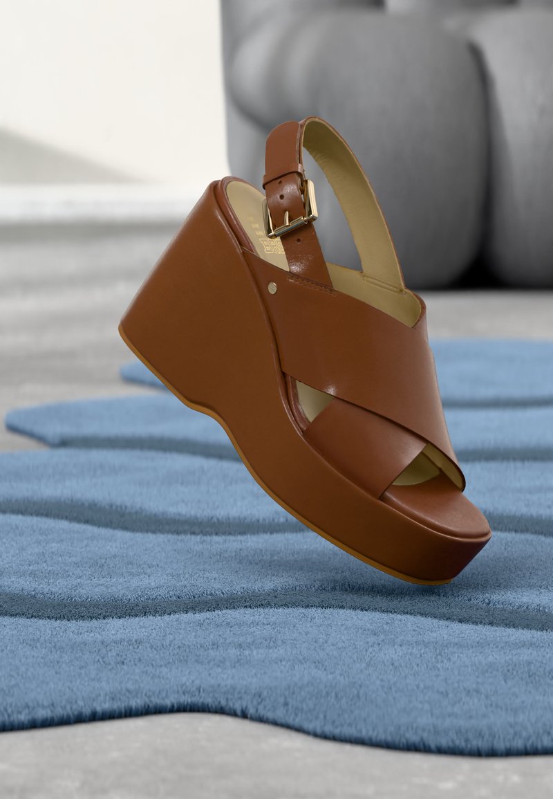 Καφετί δερμάτινο σανδάλι με χοντρό τακούνι και ρυθμιζόμενο λουράκι με αγκράφα, ακουμπισμένο σε υφασμένο μπλε χαλί εσωτερικού χώρου.