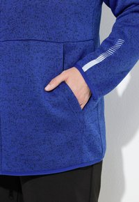 Giacca a maglia blu con superficie texture, una tasca frontale e un dettaglio bianco sulle maniche. I polsini sono rifiniti con un blu uniforme.