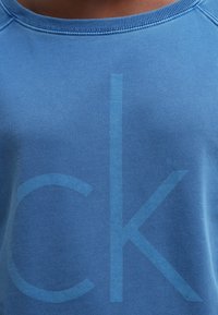 Modrá bavlněná mikina s velkými překrývajícími se písmeny v světlejším odstínu. Kulatý výstřih a jemná textura viditelná na látce.