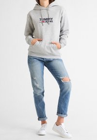 Felpa grigia con logo frontale, tasca a marsupio e cappuccio con cordino, abbinata a jeans chiari strappati e scarpe sportive bianche.