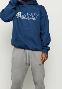Felpa con cappuccio navy di taglio over, tasca frontale e logo stampato "MLTPLY KNOWING THE STREETS" in bianco e blu. Abbinata a pantaloni della tuta grigi.