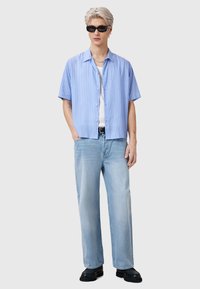 Camicia a maniche corte azzurra chiara e bianca a righe, indossata sopra una canottiera bianca, abbinata a jeans a gamba larga azzurri e scarpe nere.