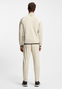 Beige athletic tracksuit made of smooth fabric, featuring a zip-up jacket and tapered pants with black accents, worn with dark sneakers.