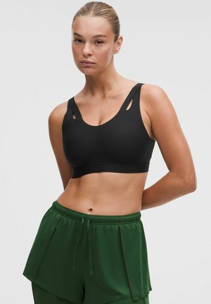 Kvinde med tilbagelagt hår iført en sort sports-bh og løstsiddende grønne atletikshorts, stående med hænderne bag ryggen.