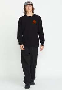 Schwarzes Langarmshirt mit orangefarbenem Logo und Grafik auf der Vorderseite. Kombiniert mit schwarzen Hosen und schwarzen Sneakers. Das Modell trägt eine Camouflage-Mütze.