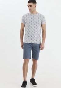 Lys grå t-shirt med et lille palmetræ mønster, parret med blå shorts. Model iført sorte sneakers, stående mod en hvid baggrund.
