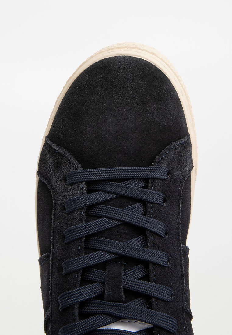 Granatowe zamszowe sneakersy charakteryzują się teksturowaną cholewką, płaskimi sznurówkami oraz kremową gumową podeszwą. W projekcie uwzględniono oczka na sznurówki oraz szczegóły w postaci przeszyć.