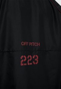 Sort vandtæt jakke med "OFF PITCH" tekst og nummeret "223" i rød, med en glat overflade og et minimalistisk design.