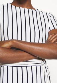 Vit klänning med tunna marinblå vertikala ränder, med båturringning och korta ärmar. Midjan knyts med ett matchande tygbälte.