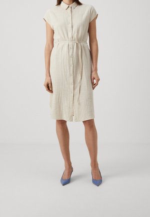 Kobieta ubrana w beżową sukienkę sięgającą do kolan, z krótkim rękawem, zapinaną na guziki, z paskiem przewiązanym w talii, oraz niebieskie szpilki z ostrym noskiem, stojąca na jednolitym tle.