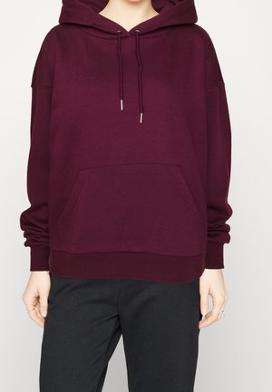 Personne portant un sweat à capuche ample violet foncé avec poche avant et cordons, associé à un pantalon noir, mains détendues de chaque côté.