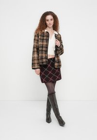 Καρό μάλλινο σακάκι σε καφέ και μαύρο, λευκό cropped πλεκτό τοπ, μίνι φούστα σε μπορντό καρό, και σκούρα μπότες μέχρι το γόνατο. Ομαλές υφές σε όλο το σύνολο.