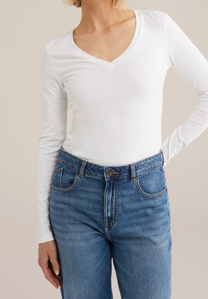 Nainen pukeutuneena valkoiseen pitkähihainen V-kaulaiseen paitaan, joka on tungettu korkeavyötäröisten sinisten farkkujen sisään, seisoo käsi lanteilla.