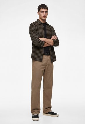 Junger Mann steht mit verschränkten Armen, trägt ein braun kariertes Hemd, schwarzes T-Shirt, beige Hose und schwarze Sneakers vor weißem Hintergrund.