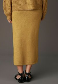 Gebreide mosterdgouden midirok met subtiele glans. Zwarte enkellaarsjes met gespen maken de look compleet.