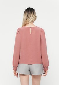 Rustfarget bluse med lange, puffete ermer, teksturert stoff og en nøkkelhullsdetalj bak, parret med grå shorts.