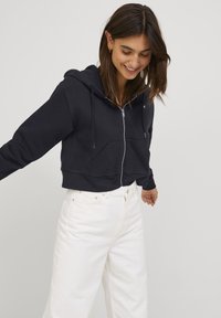 Sweatshirt preto cropped com fecho de zíper, capuz ajustável com cordão e bolsos frontais. Combinado com jeans brancos de perna larga.