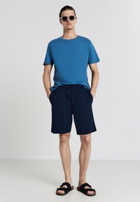 T-shirt in cotone blu, pantaloni corti navy con coulisse e sandali neri con dettaglio a fiocco. Modella che indossa occhiali da sole, in piedi con le mani in tasca.