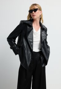 Donna che indossa una giacca di pelle nera, una blusa bianca drappeggiata, pantaloni larghi neri, occhiali da sole a occhio di gatto e orecchini a cerchio argentati su uno sfondo bianco.