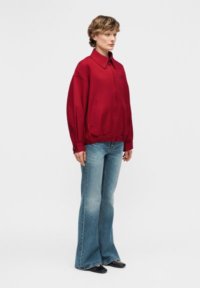 Γυναίκα με κοντά μαλλιά, που φοράει μια φαρδιά κόκκινη ζακέτα, ανοιχτό μπλε καμπάνα τζιν και μαύρα παπούτσια, όρθια μπροστά σε απλό φόντο.