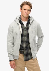 Grijze zip-up hoodie met trekkoord, gedragen over een donker geruit button-up shirt. Licht textuur, casual pasvorm, met een kangoeroezak functie.