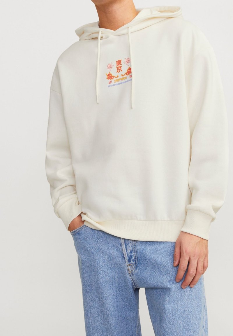 Krämfärgad hoodie med ett framsidesgrafik i rött och blått. Har en dragsko på huvan och ribbade ärmslut, tillsammans med ljusblå jeans.