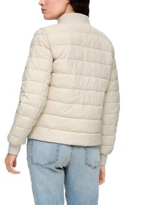 Doudoune beige avec un design matelassé, des poignets côtelés et un col montant. Portée par-dessus un jean bleu clair, mettant en avant un style minimaliste.