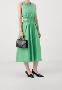 Vestido verde sin mangas con cintura elástica y detalle recortado, combinado con un bolso negro texturizado y tacones negros de punta.