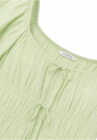 Blouse en tissu vert clair froissé avec un col carré, deux cordons à nouer à l'avant, et une étiquette de marque "OH APRIL" visible.