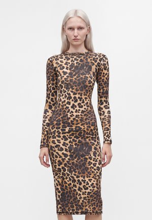 Kvinde med langt blond hår iført en tætsiddende, langærmet leopardprint kjole, stående foran en ensfarvet hvid baggrund.