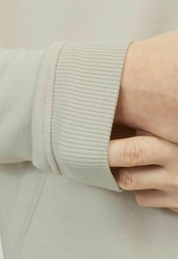 Felpa grigia chiaro con polsini a costine. Il tessuto sembra morbido e liscio, presentando un design semplice e minimal.